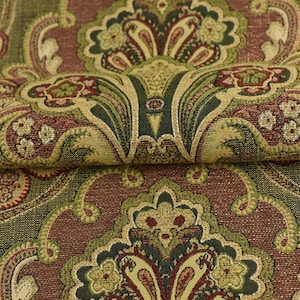 May include: Close-up of a folded fabric with a detailed paisley pattern. The design features a repeating motif in shades of olive green, burgundy, and gold. The fabric has a textured appearance, suggesting a woven material suitable for upholstery or drapery.