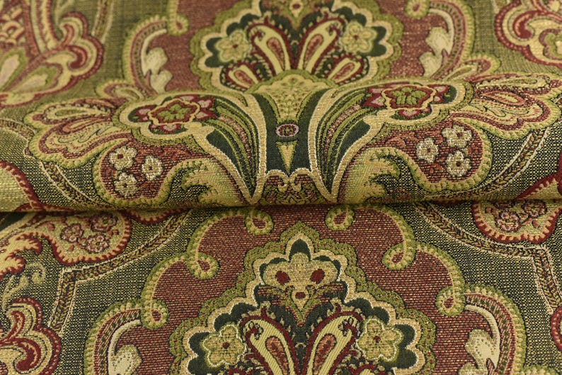 May include: Close-up of a folded fabric with a detailed paisley pattern. The design features a repeating motif in shades of olive green, burgundy, and gold. The fabric has a textured appearance, suggesting a woven material suitable for upholstery or drapery.
