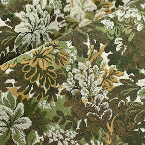 May include: Close-up of a fabric with a floral pattern. The fabric features a repeating design of stylized flowers and foliage in shades of green, brown, and cream. The fabric has a textured appearance, with visible woven threads.