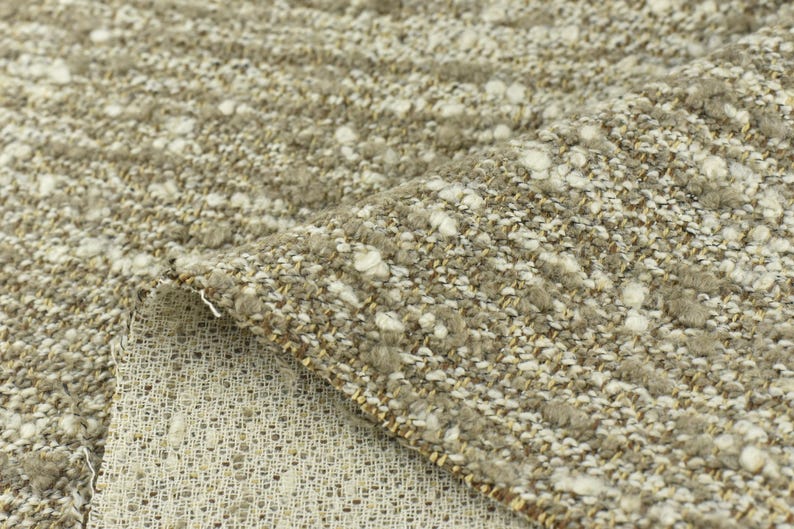 May include: Close-up of a beige and white textured fabric with a subtle sheen. The fabric has a woven appearance with small white flecks throughout.