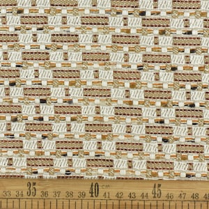 Op de afbeelding: Een close-up van een witte stof met een geweven patroon van bruine en gouden draden. De stof wordt gemeten met een liniaal die de nummers van 29 tot 53 laat zien.