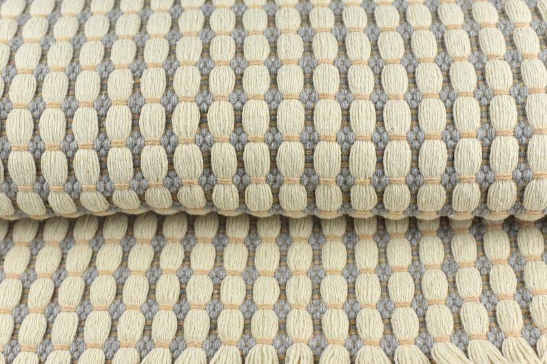 May include: Close-up of a woven fabric with a textured pattern. The fabric is a light beige color with gray and light brown accents.