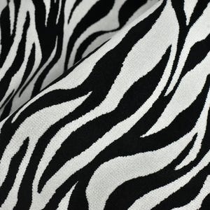 Op de afbeelding: Close-up van een stof met een gedurfd zebra streepmotief. De stof heeft zwarte strepen tegen een witte achtergrond, waardoor een contrastrijk ontwerp ontstaat. Het materiaal is gevouwen, wat de textuur en de drapering van de stof laat zien.