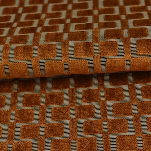 Op de afbeelding: Een gevouwen stuk stof met een gestructureerd, verhoogd geometrisch patroon in een diepe gebrande oranje kleur. De achtergrond van het textiel bestaat uit een plat, geweven grijs raster dat een contrast vormt met de zachte, pluche oranje vierkanten.