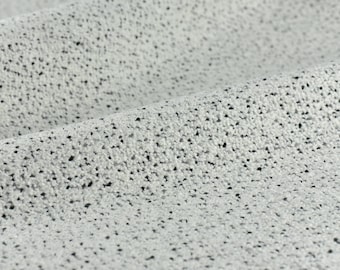 May include: Close-up of a textured fabric with a speckled pattern. The fabric is a light gray color with small, dark gray or black specks scattered across the surface. The fabric is folded, showing depth and texture.