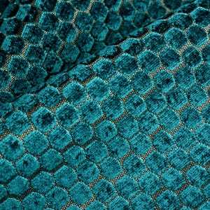 May include: Close-up of a teal fabric with a honeycomb pattern. The fabric has a plush, velvety texture and a repeating geometric design. The hexagonal shapes are slightly raised, creating a dimensional effect. The background is a neutral color.