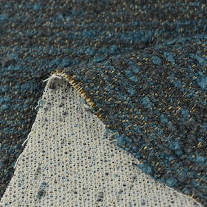 May include: Close-up of a textured fabric with a mix of dark gray, teal, and gold threads. The fabric has a nubby, woven appearance, with a lighter, open-weave backing visible where the fabric is folded over. This textile is suitable for upholstery or home decor.
