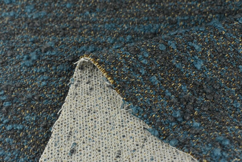 May include: Close-up of a textured fabric with a mix of dark gray, teal, and gold threads. The fabric has a nubby, woven appearance, with a lighter, open-weave backing visible where the fabric is folded over. This textile is suitable for upholstery or home decor.