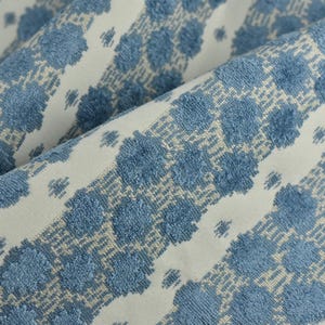Puede incluir: Primer plano de una tela doblada con un patrón repetitivo de formas florales azules texturizadas sobre un fondo crema. La tela tiene un aspecto suave y afelpado, adecuada para tapicería o fines decorativos.