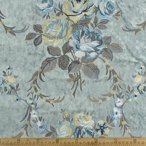 May include: Close-up of a fabric with an embroidered floral design. The pattern features large, detailed roses in shades of blue and yellow, with gray-brown leaves and stems. The background is a muted teal color. A ruler is at the bottom.