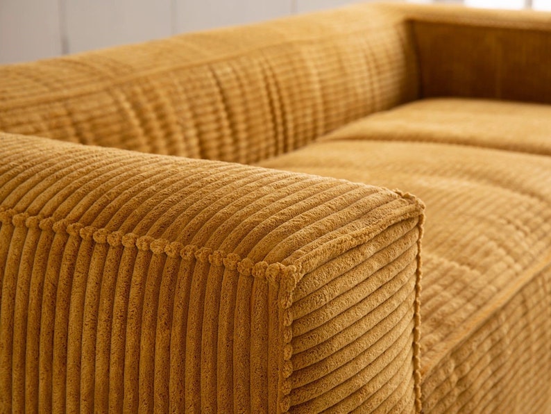 K&ouml;nnte beinhalten: Nahaufnahme eines senfgelben Cord-Sofas mit gestepptem R&uuml;cken und Armlehnen. Der Stoff hat eine gerippte Textur.