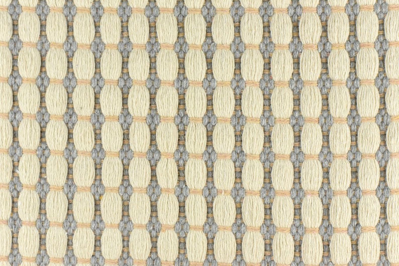 May include: A close-up of a woven fabric with a repeating pattern of small, light beige circles surrounded by a darker gray border. The fabric has a subtle texture and a slightly raised surface.