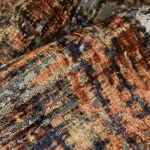 May include: A textured fabric featuring an abstract pattern with a blend of deep rust orange, dark navy blue, and metallic silver threads. The material displays a dense, woven pile with visible folds that create shadows and highlight the varied colors.