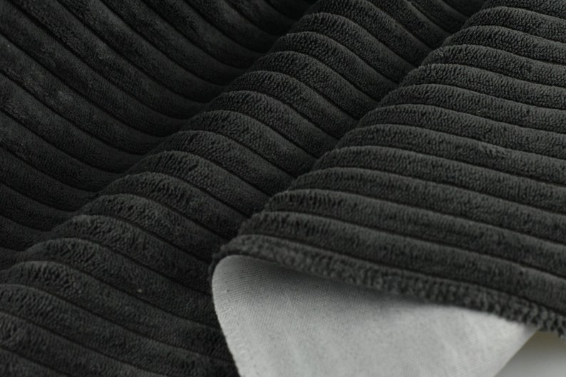 May include: Close-up of black corduroy fabric with a soft, textured surface. The fabric is folded in half, revealing a grey backing.
