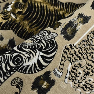 May include: Close-up of a tan fabric with a repeating animal print design. The design features tigers and leopards in black, white, and gold. The fabric is folded, showing the pattern's repeating nature.
