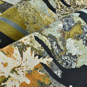 May include: Close-up of folded fabric with an abstract design. The pattern features black, white, olive green, and shades of orange and grey. The fabric has a textured appearance.