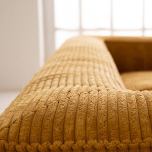 K&ouml;nnte beinhalten: Nahaufnahme eines senfgelben Cord-Sofas mit einer gesteppten Oberfl&auml;che. Das Sofa ist aus einem weichen, pl&uuml;schigen Stoff gefertigt und wirkt komfortabel und einladend.