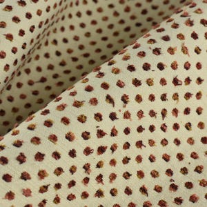 以下が含まれることがあります： 薄いクリーム色の生地のクローズアップ。小さな濃い赤色のドットが繰り返しパターンで並んでいます。