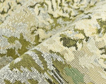 Op de afbeelding: Close-up van een textiel met een bloemenpatroon in tinten groen, crème en grijs. De stof is gevouwen en onthult de gedetailleerde weefsel en kleurvariaties. Het ontwerp heeft een vintage of botanische uitstraling.