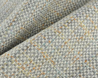 Op de afbeelding: Close-up van een textuurdoek met een geweven patroon. De stof is lichtgrijs met hints van blauw, oranje en geel.