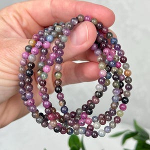 May include: A multi-strand bracelet composed of small, smooth, spherical beads in various shades of pink, purple, blue, green, and grey. The beads are strung together in a circular arrangement, showcasing a diverse range of translucent and opaque colors.
