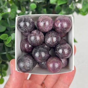 May include: A collection of small, polished, spherical stones in shades of deep purple, magenta, and dark grey. These smooth, rounded mineral spheres are packed together inside a square white cardboard box, displaying natural crystalline textures.