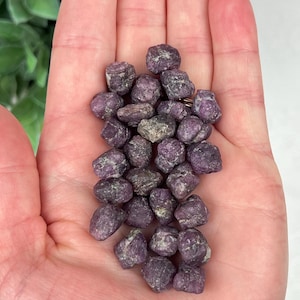 May include: A collection of small, raw, deep purple and magenta-toned mineral stones with a rough, textured surface. These irregular, pebble-like crystals feature natural variations in color and shape, clustered together in a dense pile.