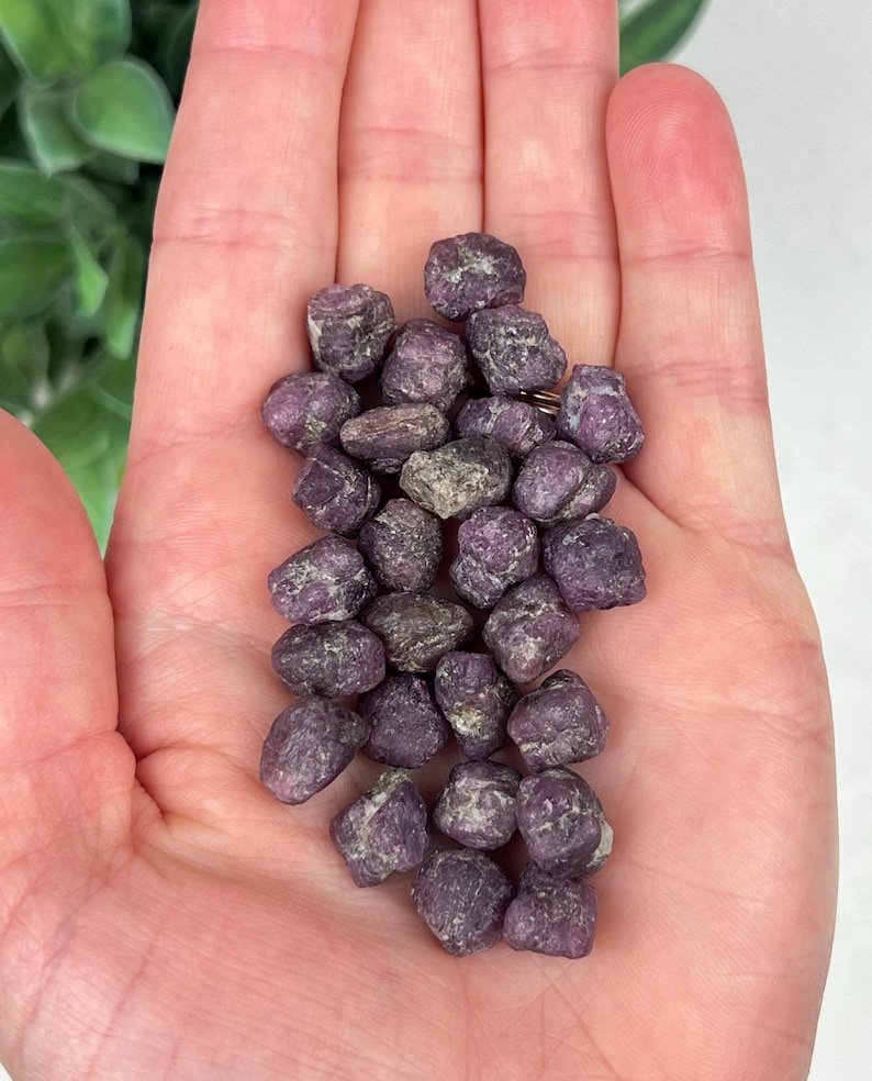 May include: A collection of small, raw, deep purple and magenta-toned mineral stones with a rough, textured surface. These irregular, pebble-like crystals feature natural variations in color and shape, clustered together in a dense pile.