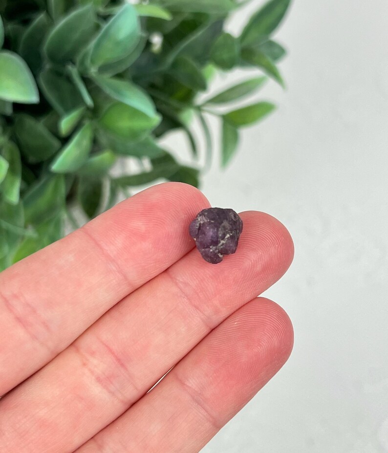 May include: A small, raw, deep purple gemstone with an irregular, multifaceted surface. The stone displays a dark, saturated violet hue with natural texture and uneven edges, showcasing a compact, unpolished form typical of a rough mineral specimen.