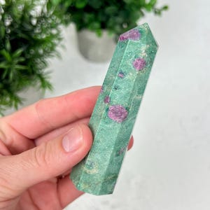 May include: A polished hexagonal crystal tower featuring a mottled green base stone with embedded vibrant magenta-pink mineral inclusions. The stone has a smooth, faceted surface that tapers to a point at the top, showcasing natural color variations.