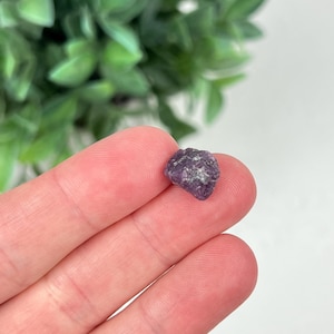 May include: A small, raw, deep purple gemstone with an irregular, multifaceted surface texture. The stone displays natural variations in color intensity and crystalline structure, appearing as a rough, unpolished mineral fragment.