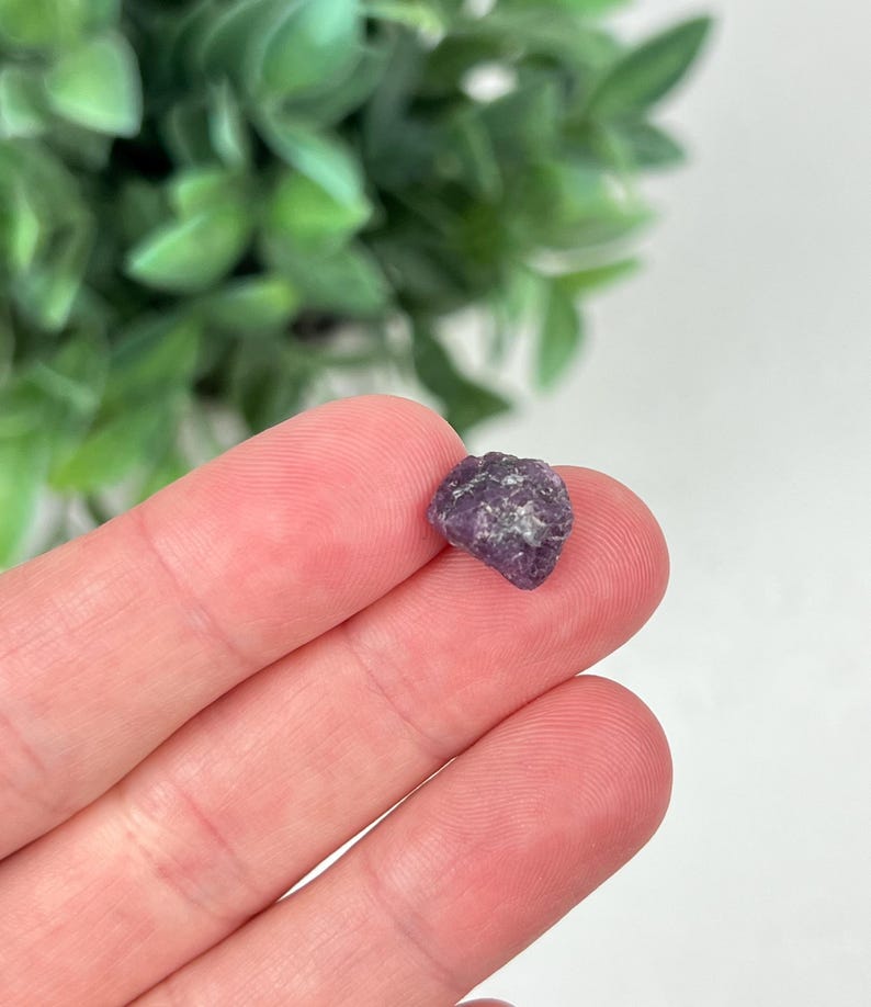 May include: A small, raw, deep purple gemstone with an irregular, multifaceted surface texture. The stone displays natural variations in color intensity and crystalline structure, appearing as a rough, unpolished mineral fragment.