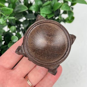 May include: A circular dark brown wooden display stand featuring a smooth, slightly concave top surface with a raised rim and four carved, angular feet extending from the base, showcasing a natural wood grain texture throughout the entire object.