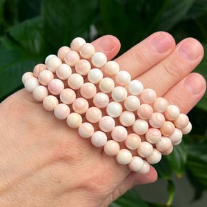 May include: Several strands of pink and white round beads, likely gemstones, are displayed on a hand. The beads have a smooth, polished appearance and vary in shades from pale pink to creamy white. The background is a blurred green.