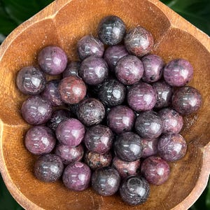 May include: A bowl filled with purple and black tumbled gemstone spheres. The spheres are smooth and polished, with a variety of sizes and shapes.