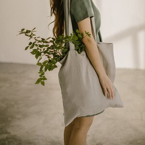 Puede incluir: Una mujer con un vestido de lino verde lleva una gran bolsa de tela gris claro con una planta verde frondosa en su interior.