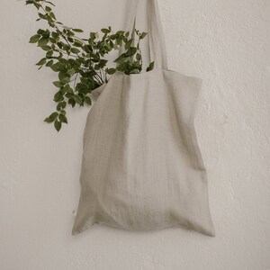 Puede incluir: Una bolsa de lona beige colgada de una pared blanca con una rama de hojas verdes y flores blancas en su interior.