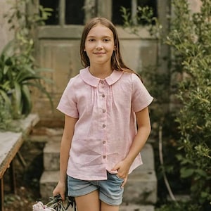 May include: A young person wearing a pink linen shirt with a Peter Pan collar and short sleeves. They are wearing denim shorts and holding a bouquet of white flowers.