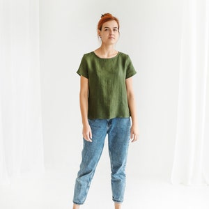 May include: A woman wearing a green linen shirt with short sleeves and blue jeans. She is standing on a white background.