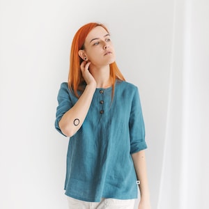 May include: A woman with red hair wearing a teal blue linen shirt with a button-up front and white pants. She has a small circle tattoo on her left arm.