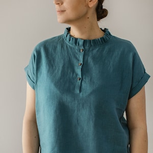 May include: A teal linen shirt with a ruffled collar and a single button closure. The shirt has short sleeves and a relaxed fit.