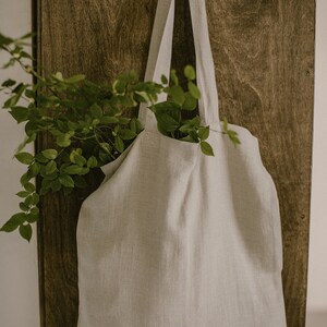 Puede incluir: Una bolsa de lona de lino gris claro con una textura de tejido natural colgada de una puerta de madera. La bolsa tiene dos correas largas y est&aacute; vac&iacute;a.