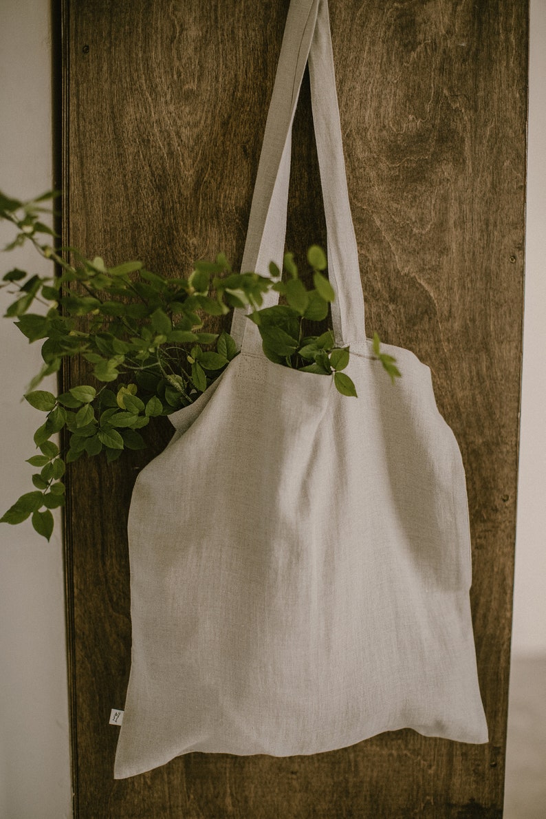 Puede incluir: Una bolsa de lona de lino gris claro con una textura de tejido natural colgada de una puerta de madera. La bolsa tiene dos correas largas y est&aacute; vac&iacute;a.