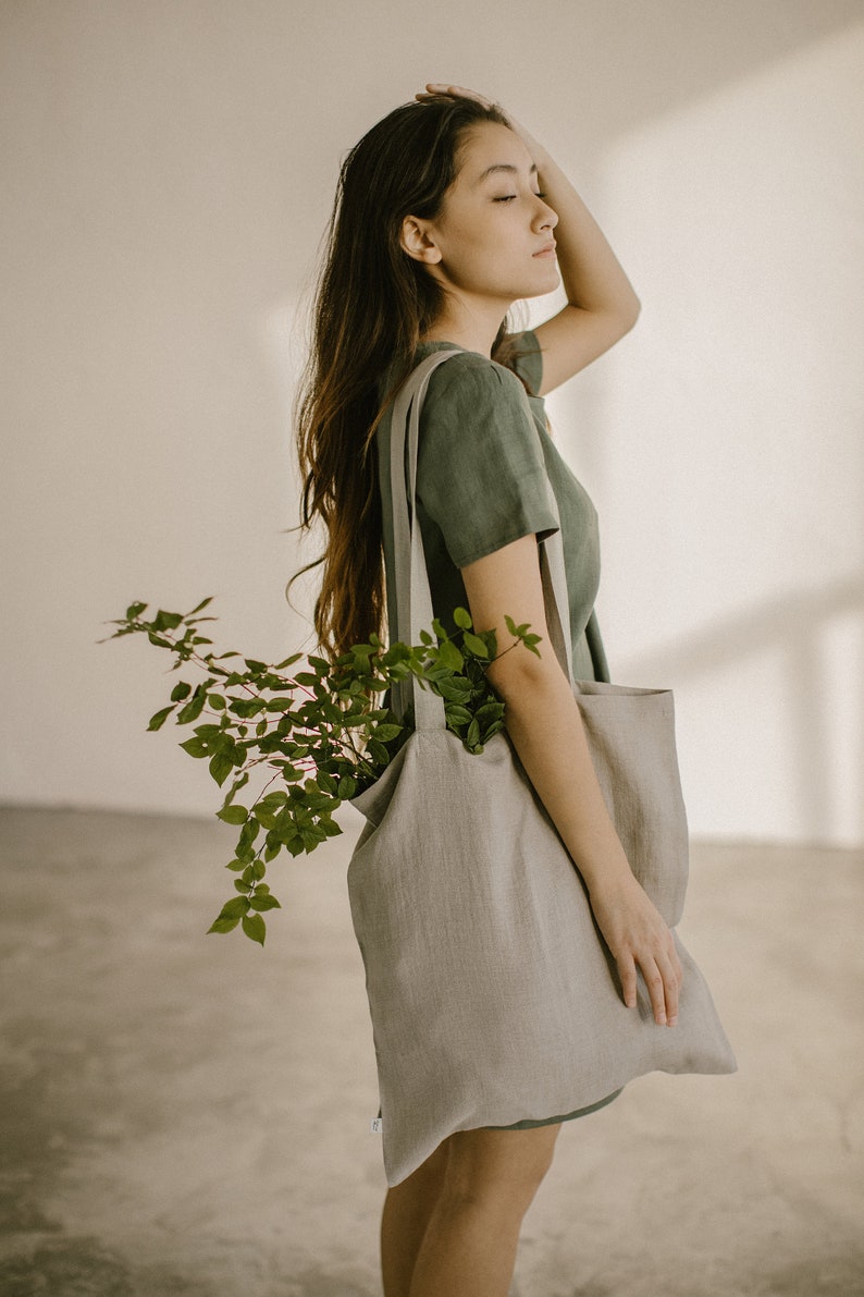 Puede incluir: Una mujer con un vestido de lino verde lleva una gran bolsa de tela gris claro con una planta verde frondosa en su interior.