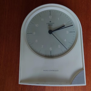 Puede incluir: Reloj analógico blanco con agujas negras y una esfera plateada. La esfera del reloj tiene números negros y el texto "JUNGHANS MEGA" impreso en ella. El reloj está controlado por radio y tiene un marco blanco con un acento gris en la esquina inferior derecha.
