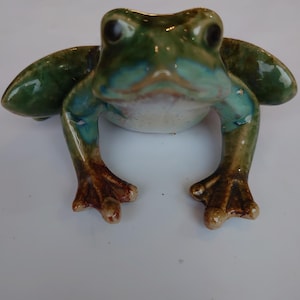 Könnte beinhalten: Eine Keramik-Froschfigur mit glänzendem Finish. Der Frosch ist hauptsächlich grün mit blauen und braunen Akzenten. Die Figur hat große Augen und ausgestreckte Arme, was ihr ein skurriles Aussehen verleiht. Die Füße des Frosches sind warmbraun.