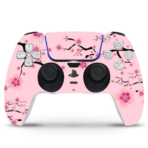 May include: A pink PlayStation 5 DualSense controller featuring a cherry blossom design. The controller has black buttons and analogue sticks.