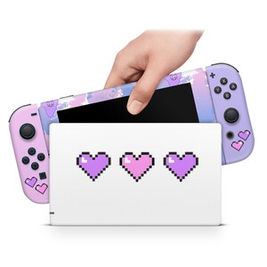 May include: A pink and purple Nintendo Switch console with a pixelated heart design. The console is closed and in a white case with three pixelated hearts on the front.