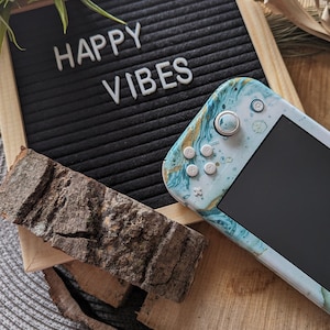 May include: A black letter board with the words "HAPPY VIBES" displayed. A teal and gold Nintendo Switch console with a marble design is on a wooden surface next to the letter board.
