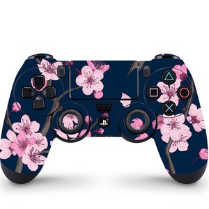 Puede incluir: Un mando de PlayStation 4 negro con un diseño floral con flores de cerezo rosas sobre un fondo azul oscuro.